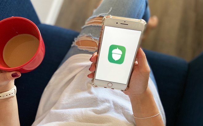 A Person Holding a Drink and a Phone with Acorns Logo on the Background
