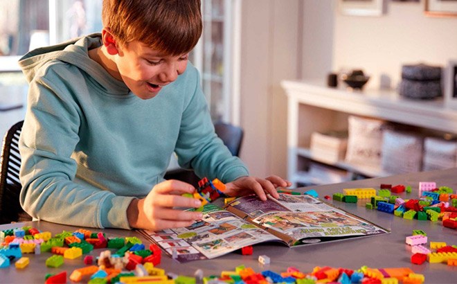 A Smiling Boy Reading the LEGO Magazine at a Desk With Lots of LEGOs Around the Magazine