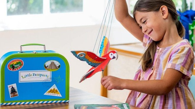 Little Girl Playing With a Parrot Toy Next to a Little Passports Suitcase Box