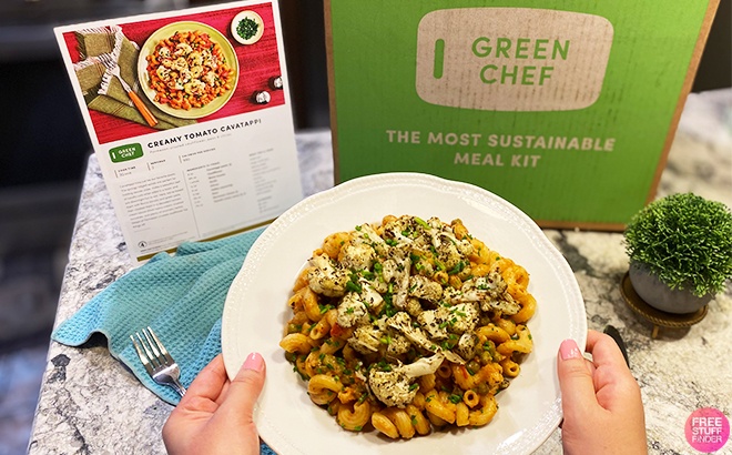 Person Holding a Plate with Pasta Meal on a Table Next to Green Chef Box