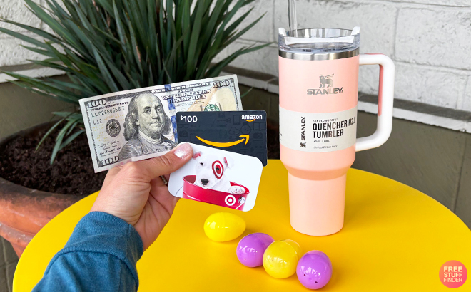 A Person Holding a 100 Dollar Bill a Target and Amazon Gift Card with a Stanley Peach Quencher Tumnler and Easter Eggs in the Background
