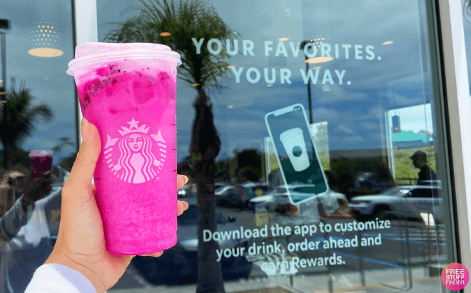 Hand holding Starbucks pink dragon drink infront of Starbucks store with rewards app information