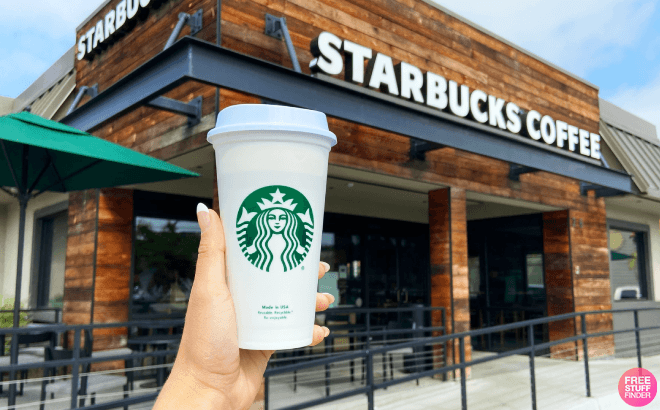 Hand holding white reusable Starbucks cup in front of a café