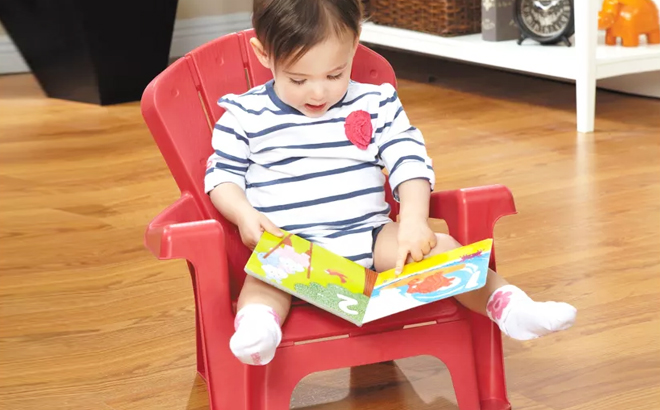 A Toddler Reading a Book and Sitting on Red Little Tikes Garden Outdoor Portable Chair