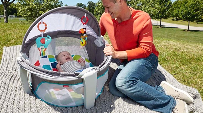 Baby Laughing in Fisher Price Portable Bassinet and Play Space with the Dad Sitting Next to it on a Picnic Blanket