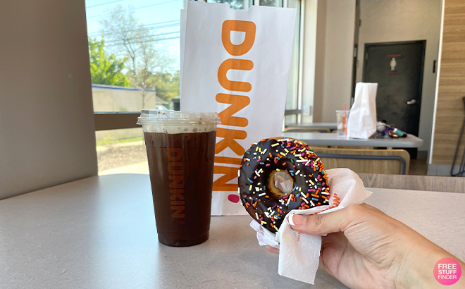 A Hand Holding a Donut Next to a Dunkin Donuts Coffee and Takeout Bag