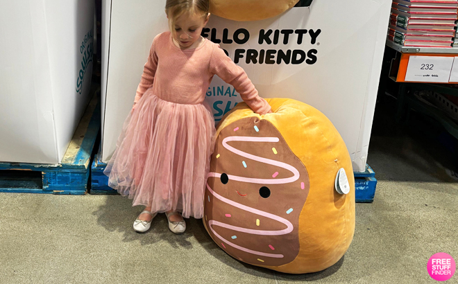 Girl in Pink Dress Standing Next to Squishmallows Deja The Chocolate Frosted Donut Jumbo Plush Toy