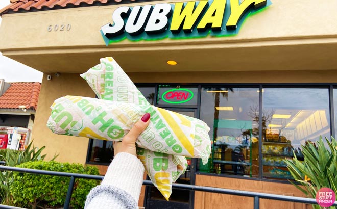 a Hand Holding Two Footlong Subs in Front of a Subway Chain