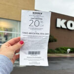 Hand Holding Kohls Coupons for 20 off In Front of Kohls Store Sign