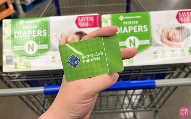 A Hand Holding a Sam's Club Membership Card with Two Boxers of Diapers in the Background