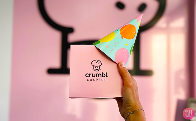 A Person Holding a Cumbl Cookie Box with a Party Hat on in Front of the Crumbl Sign Inside the Store