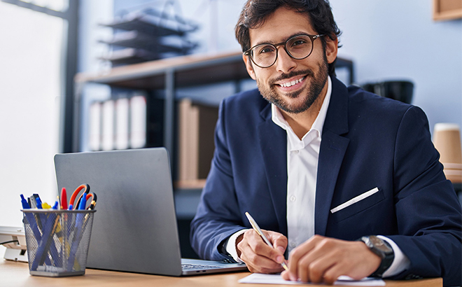 A Person Holding a Pen and Paper Sitting at a Desk with a Computer in Front of Them