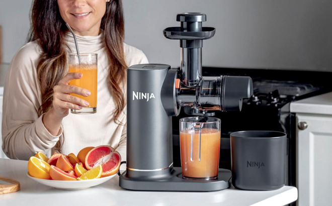 A Woman Drinking Fresh Juice next to the Ninja NeverClog Cold Press Juicer