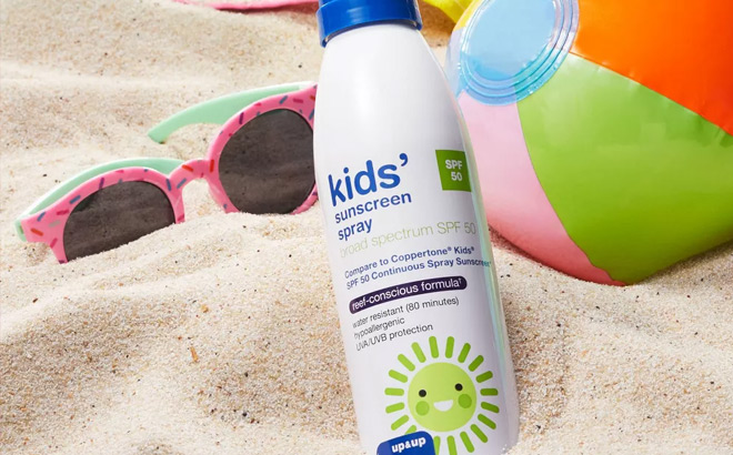 Up Up SPF 50 Kids Sunscreen Spray on a Beach Next to a Pair of Sunglasses and a Beach Ball
