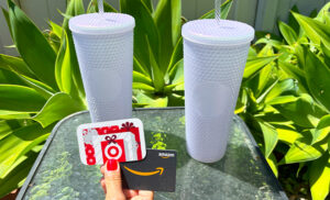 A Person Holding a Target and Amazon Gift Card with Two Lilac Starbucks Tumblers in the Background on a Table