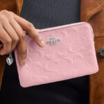 A Woman Holding Coach Outlet Corner Zip Wristlet In Signature Leather With Heart Charm